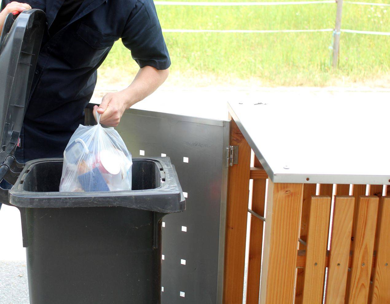 Person wirft Muellbeutel in schwarze Tonne vor Muelltonnenbox Holz mit Edelstahlklappe