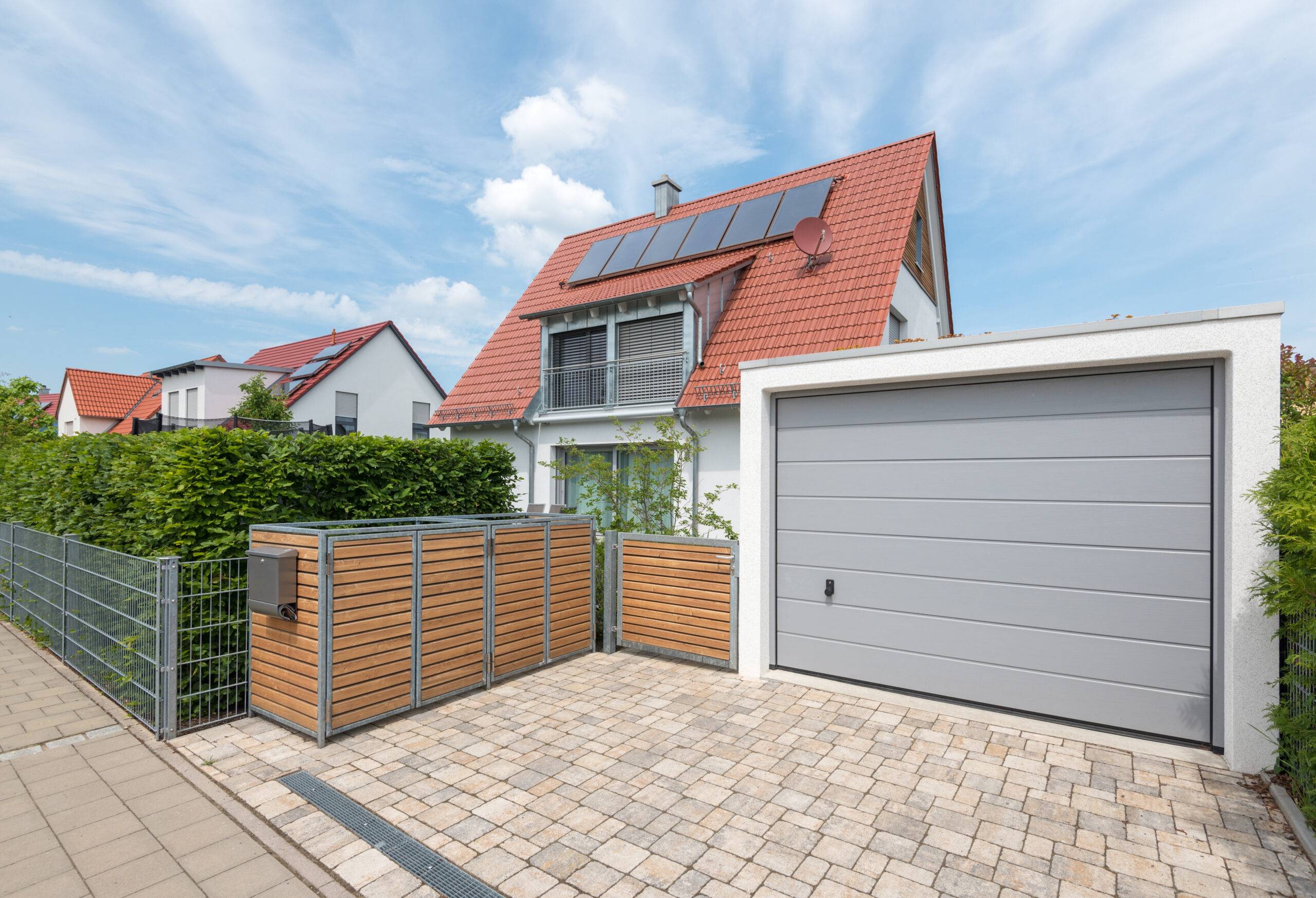 Muelltonnenbox Holz mit Metallrahmen auf gepflastertem Vorplatz neben Garage